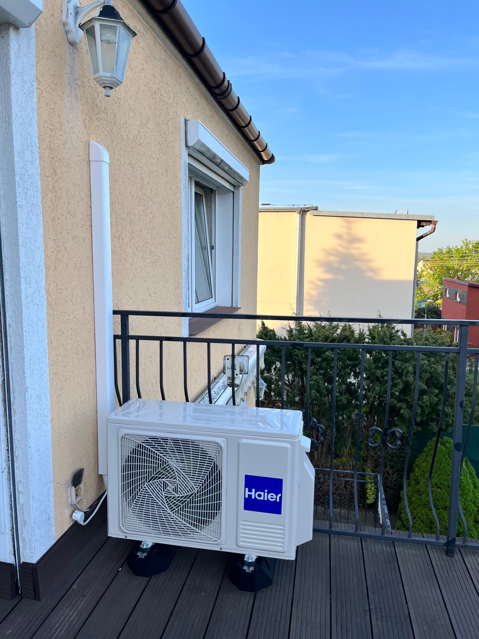 Biały klimatyzator Haier zamontowany na balkonie z drewnianą podłogą, widoczne elementy instalacji na elewacji budynku, czarne barierki i zieleń w tle.