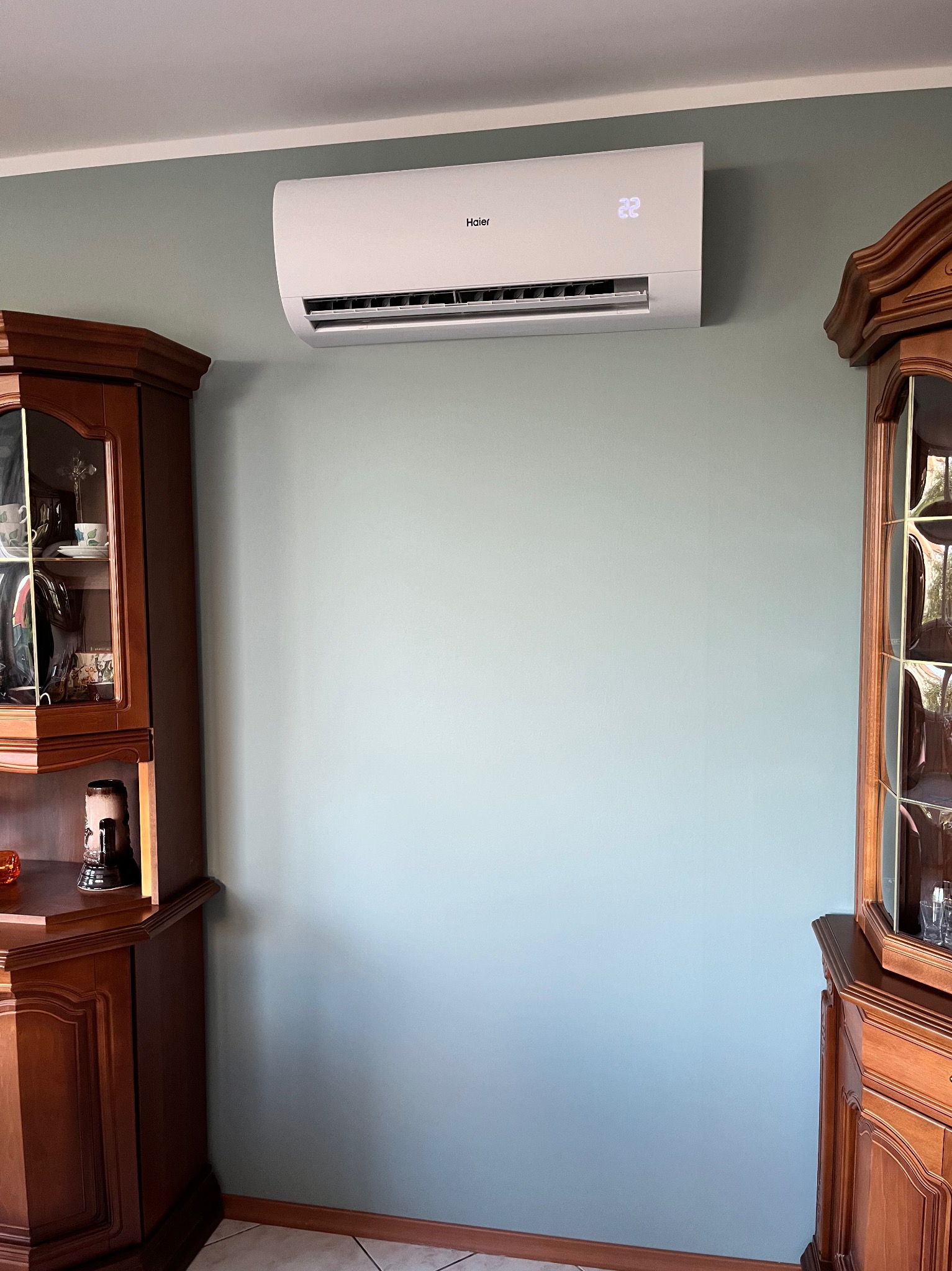 Ścienna klimatyzacja Haier w jasnym wnętrzu, wyświetlająca temperaturę 22 stopnie, zamontowana na tle pastelowej ściany między dwoma drewnianymi kredensami.