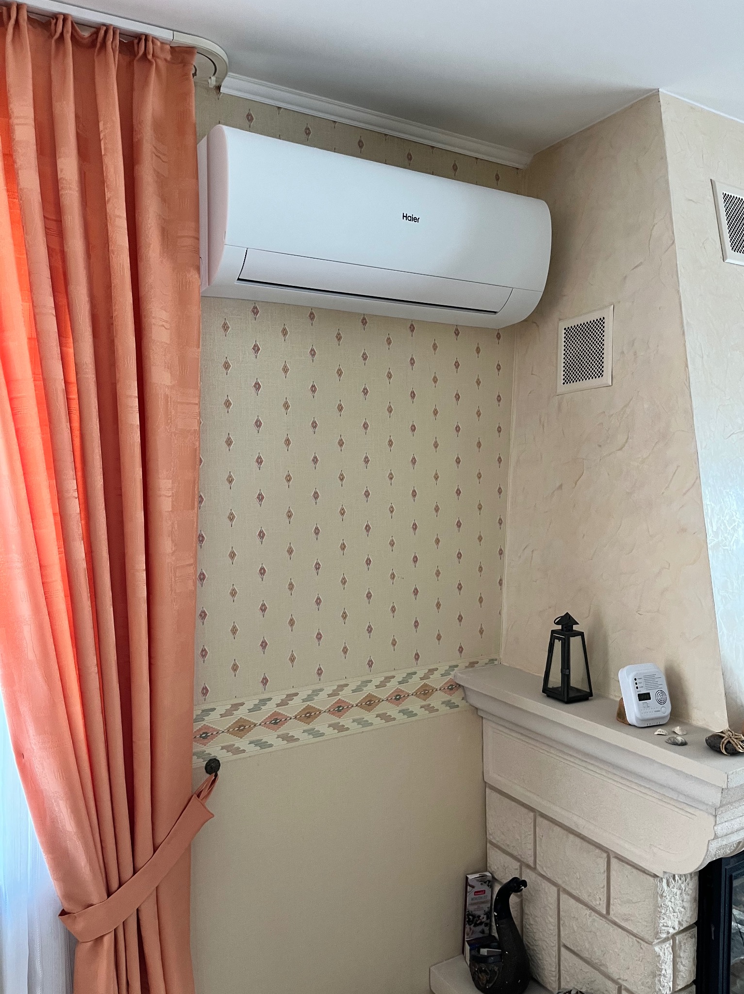Biały klimatyzator Haier zamontowany na ścianie z wzorzystą tapetą w pokoju z beżowymi zasłonami i kominkiem. Widoczne kratki wentylacyjne.