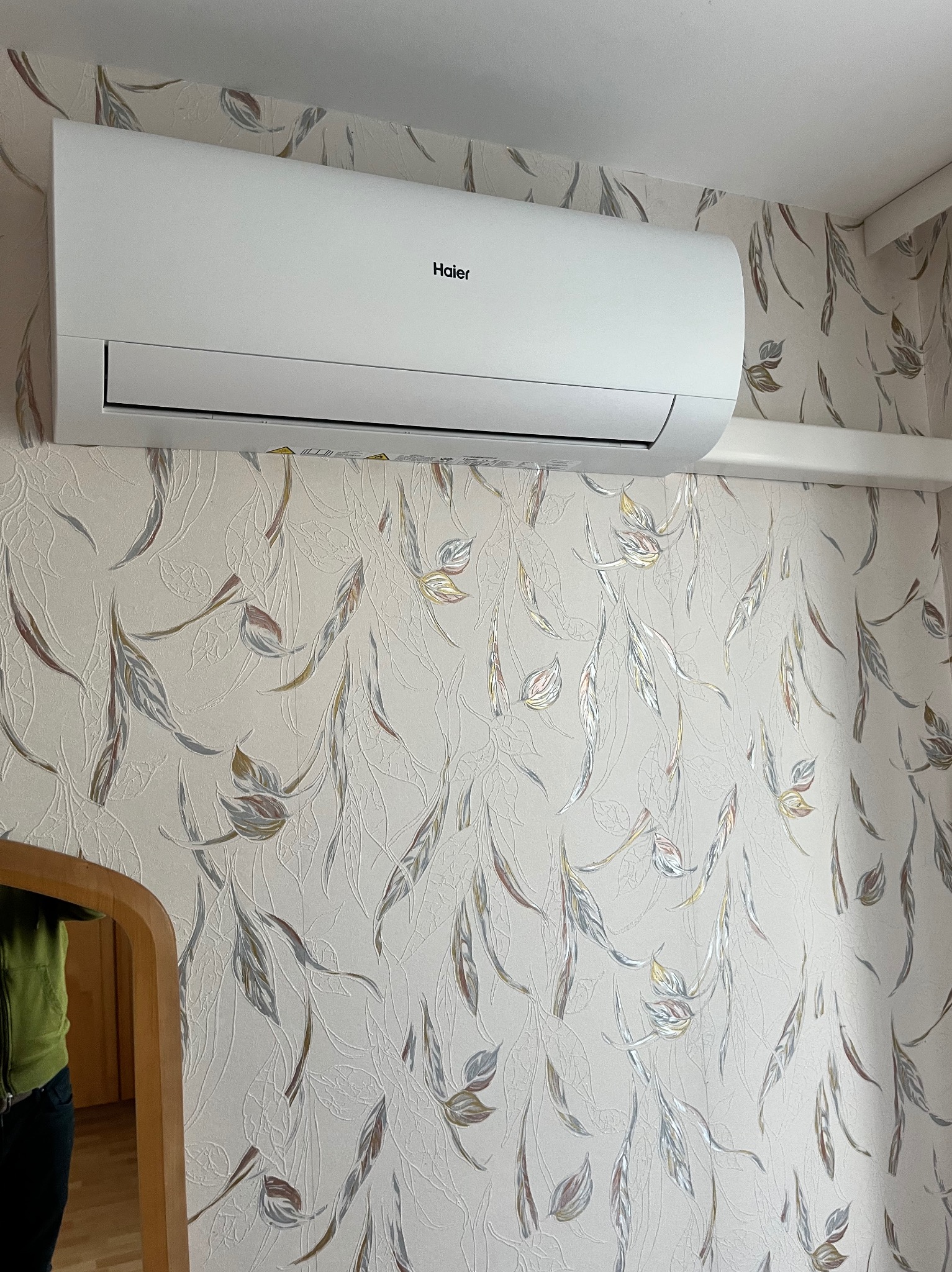 Biały klimatyzator Haier zamontowany na ścianie z tapetą w liście. Widoczne odbicie w lustrze. Ujęcie z dołu, podkreśla estetykę urządzenia i dopasowanie do wnętrza.