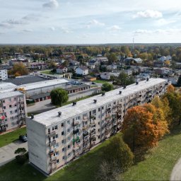 Nieruchomości Angelika Kawczyńska - Panoramiczny widok z lotu ptaka na osiedle mieszkaniowe z blokami z balkonami, otoczone zielenią i drzewami w jesiennych barwach, w tle zabudowa jednorodzinna i las.