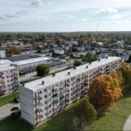 Panoramiczny widok z lotu ptaka na osiedle mieszkaniowe z blokami z balkonami, otoczone zielenią i drzewami w jesiennych barwach, w tle zabudowa jednorodzinna i las.