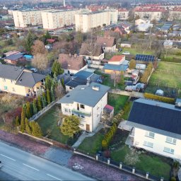 Nieruchomości Angelika Kawczyńska - Widok z lotu ptaka na osiedle domów jednorodzinnych i bloków mieszkalnych, z panelami słonecznymi na dachach niektórych budynków i samochodem osobowym na ulicy.