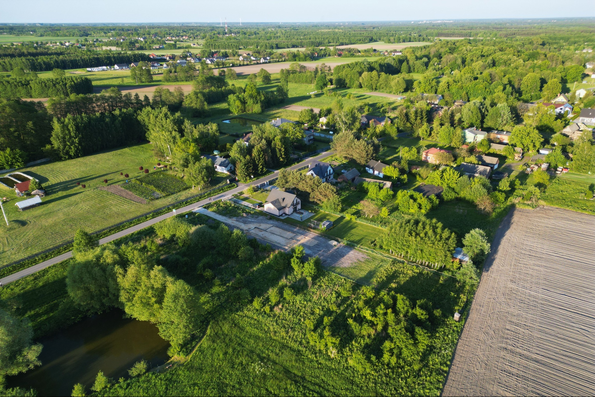 Panoramiczny widok z drona na wiejską okolicę z domami jednorodzinnymi, polami uprawnymi i lasami w letnim świetle.