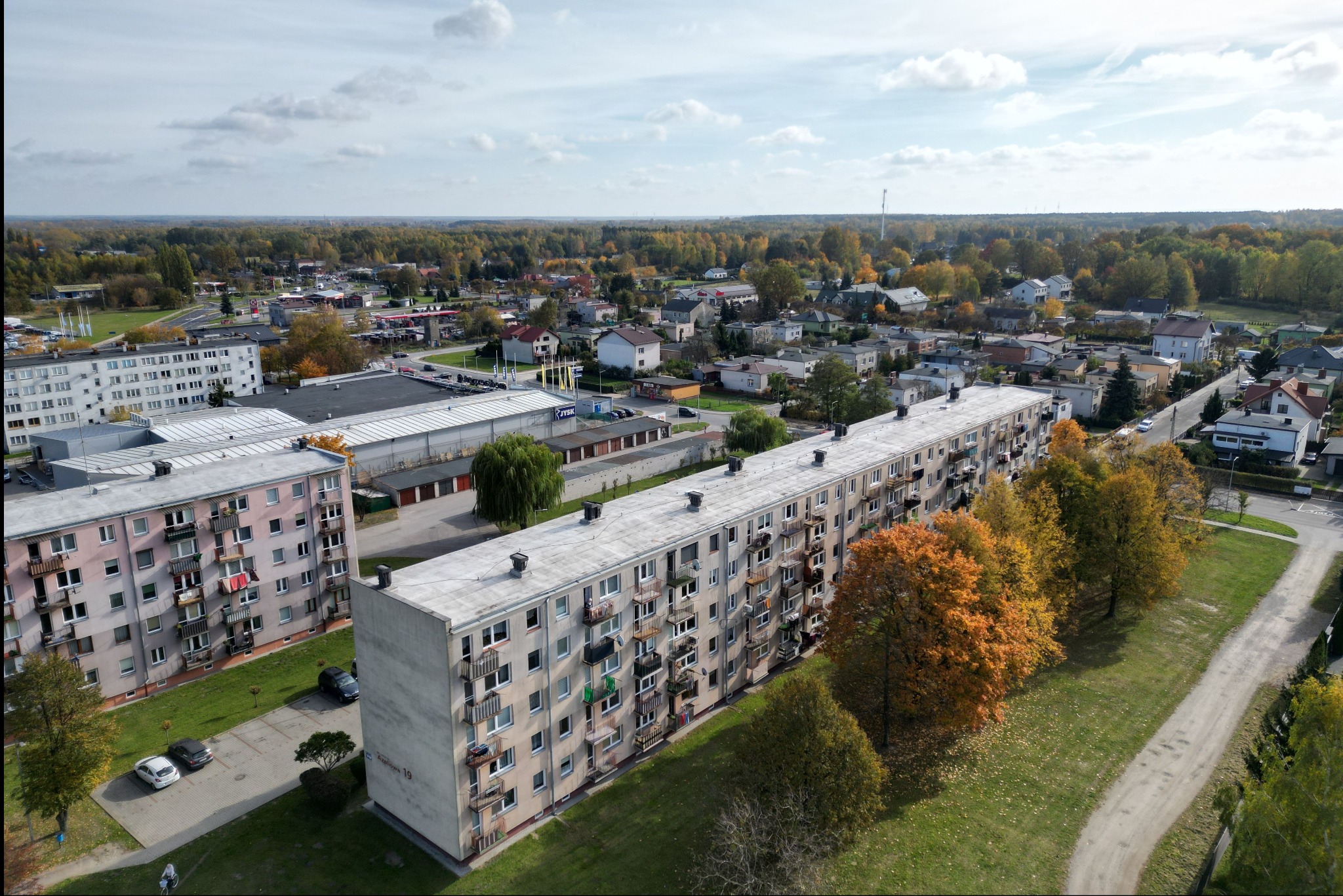 Panoramiczny widok z lotu ptaka na osiedle mieszkaniowe z blokami z balkonami, otoczone zielenią i drzewami w jesiennych barwach, w tle zabudowa jednorodzinna i las.