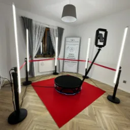 Oświetlona, obracająca się platforma do zdjęć 360 stopni na czerwonym dywanie, otoczona barierką i lampami LED w pomieszczeniu z plakatem reklamowym.