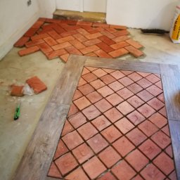 Przedsiębiorstwo Budowlane - Układanie ceglanych płytek podłogowych w ramie z drewnopodobnych paneli, widoczne narzędzie do cięcia i worek z klejem w tle.