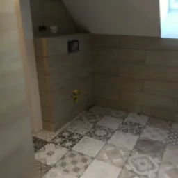 Ukończona łazienka na poddaszu: beżowe płytki imitujące drewno na ścianach, mozaika patchworkowa na podłodze, widoczne przyłącza do armatury.