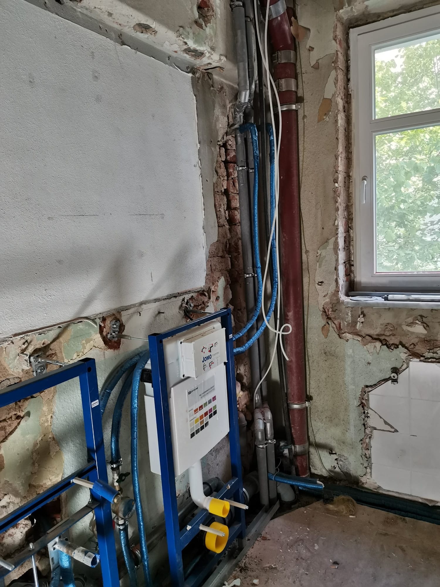 Przygotowanie do montażu podtynkowego stelaża WC w surowym stanie, widoczne rury instalacyjne i fragmenty starej glazury na ścianie, okno w tle.