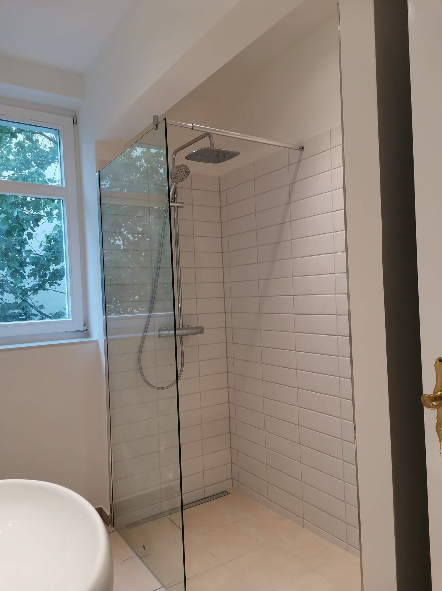 Wykończona kabina prysznicowa typu walk-in z odpływem liniowym, obłożona białymi, poziomymi płytkami w Bolesławcu. Widoczne okno z widokiem na zieleń i fragment umywalki.