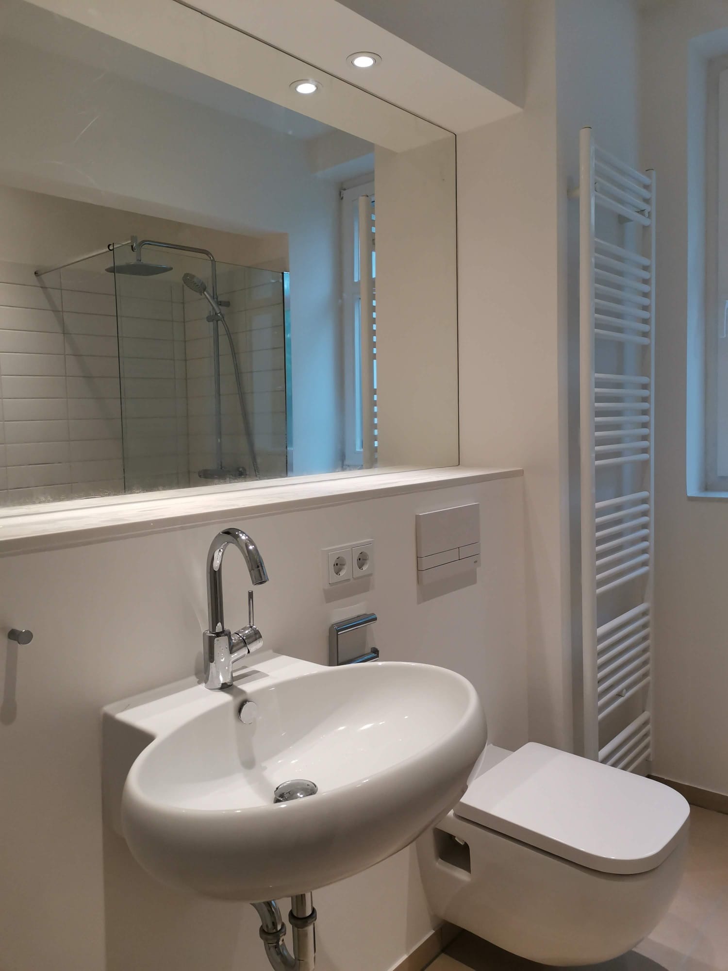 Nowoczesna, minimalistyczna łazienka z owalną umywalką nablatową, chromowaną baterią, podwieszaną toaletą i grzejnikiem drabinkowym w kolorze białym. W lustrze odbija się kabina prysznicowa...