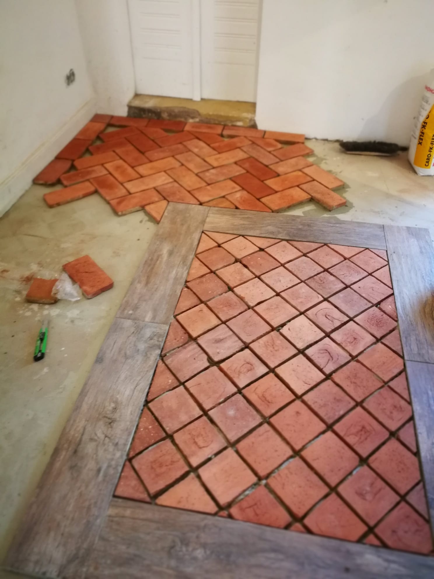 Układanie ceglanych płytek podłogowych w ramie z drewnopodobnych paneli, widoczne narzędzie do cięcia i worek z klejem w tle.