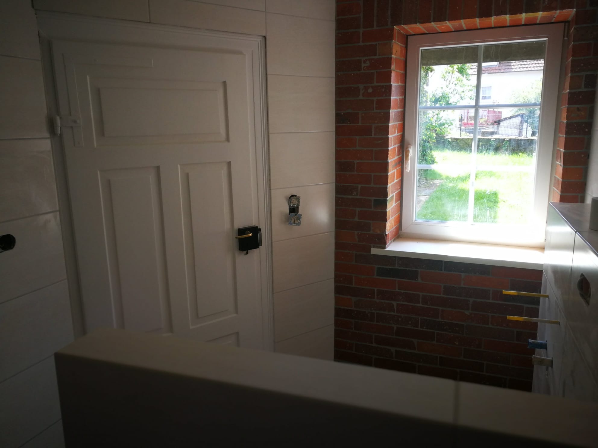 Wykończona łazienka z białą, panelową ścianą, białymi drzwiami i oknem w czerwonej ceglanej ramie z widokiem na zieleń.