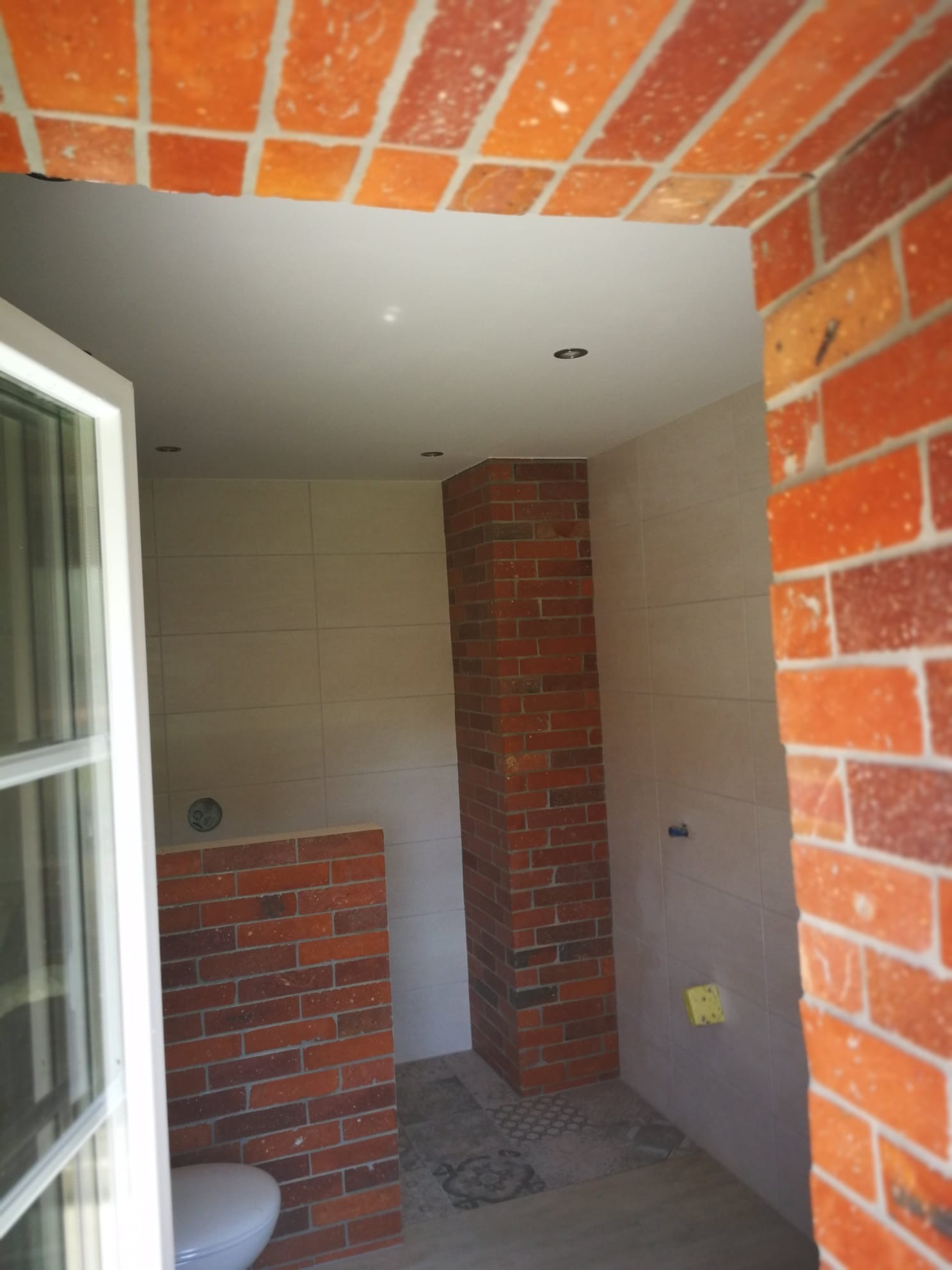 Wnętrze łazienki w trakcie wykańczania, widoczne płytki na ścianach, element dekoracyjny z cegły, częściowo zamontowana toaleta oraz otwór drzwiowy z widocznym fragmentem ceglanej konstrukcji.