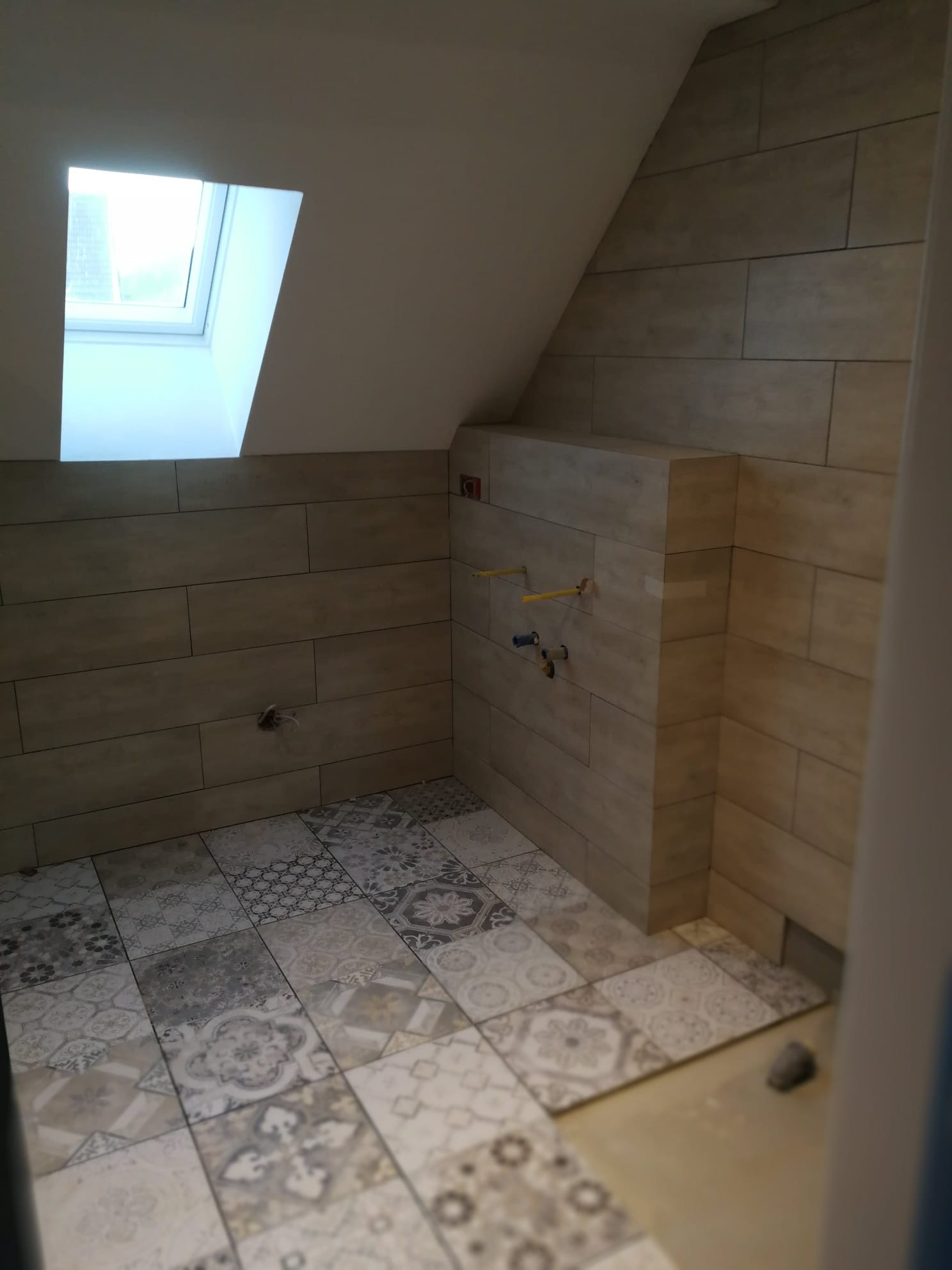 Ukończona łazienka na poddaszu z beżowymi płytkami imitującymi drewno na ścianach, wzorzystą patchworkową podłogą i oknem dachowym; widoczne wystające rury instalacji wodnej.