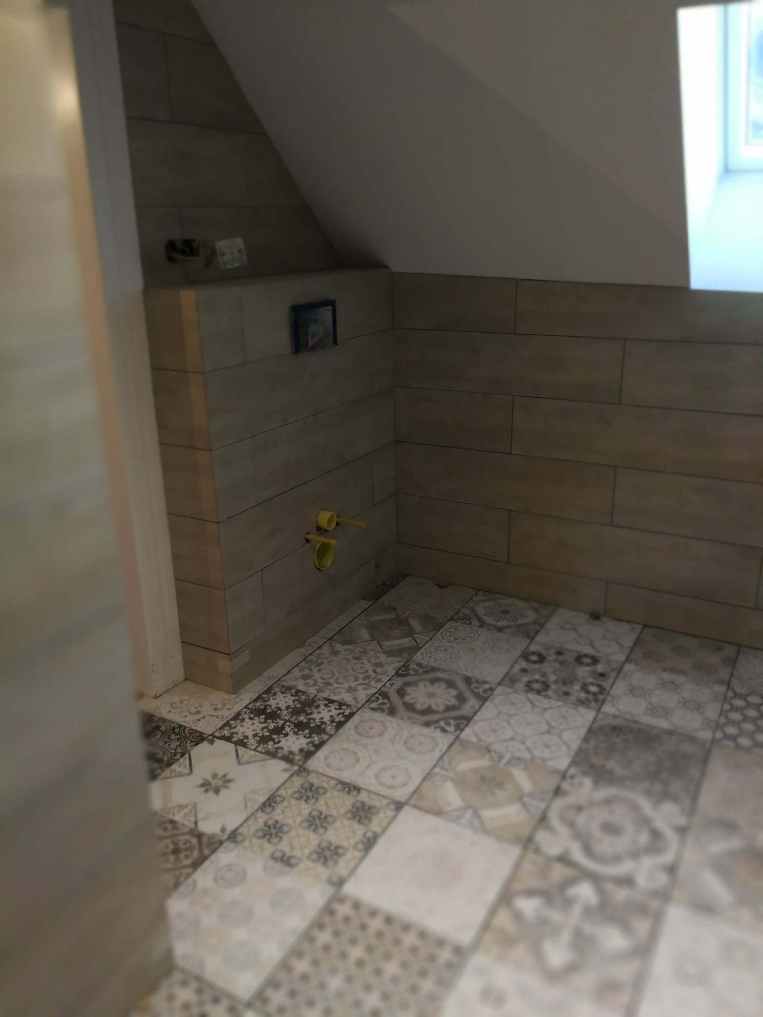 Ukończona łazienka na poddaszu: beżowe płytki imitujące drewno na ścianach, mozaika patchworkowa na podłodze, widoczne przyłącza do armatury.