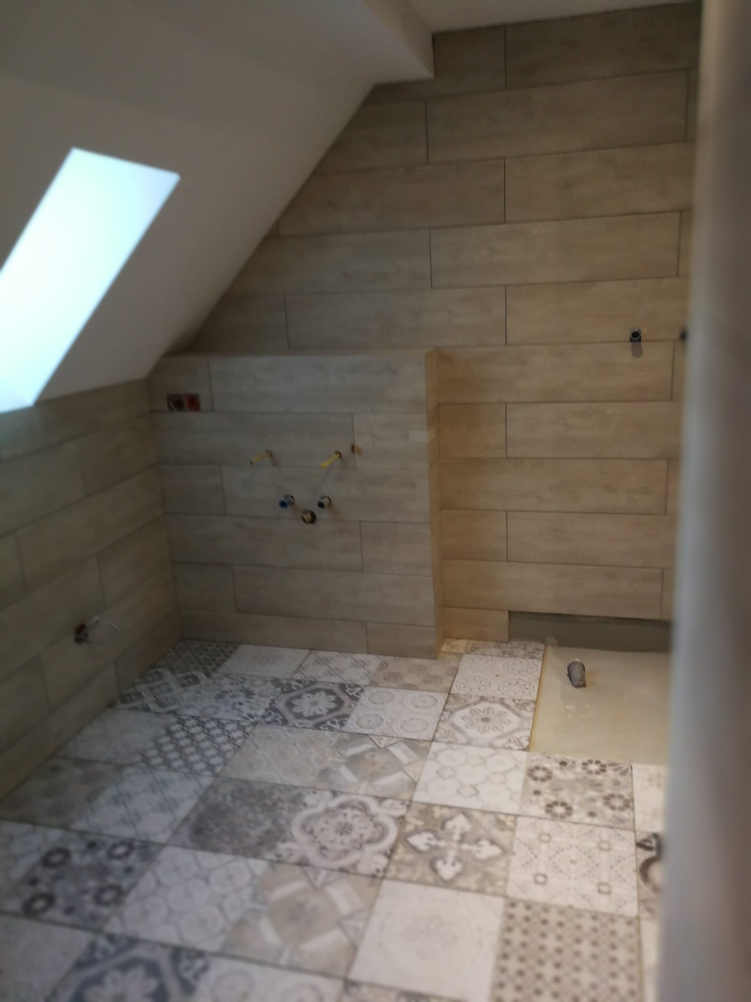 Wykończona łazienka na poddaszu z ułożonymi płytkami podłogowymi o różnych wzorach i beżowymi płytkami imitującymi drewno na ścianach, widoczne wystające rury instalacji wodnej.