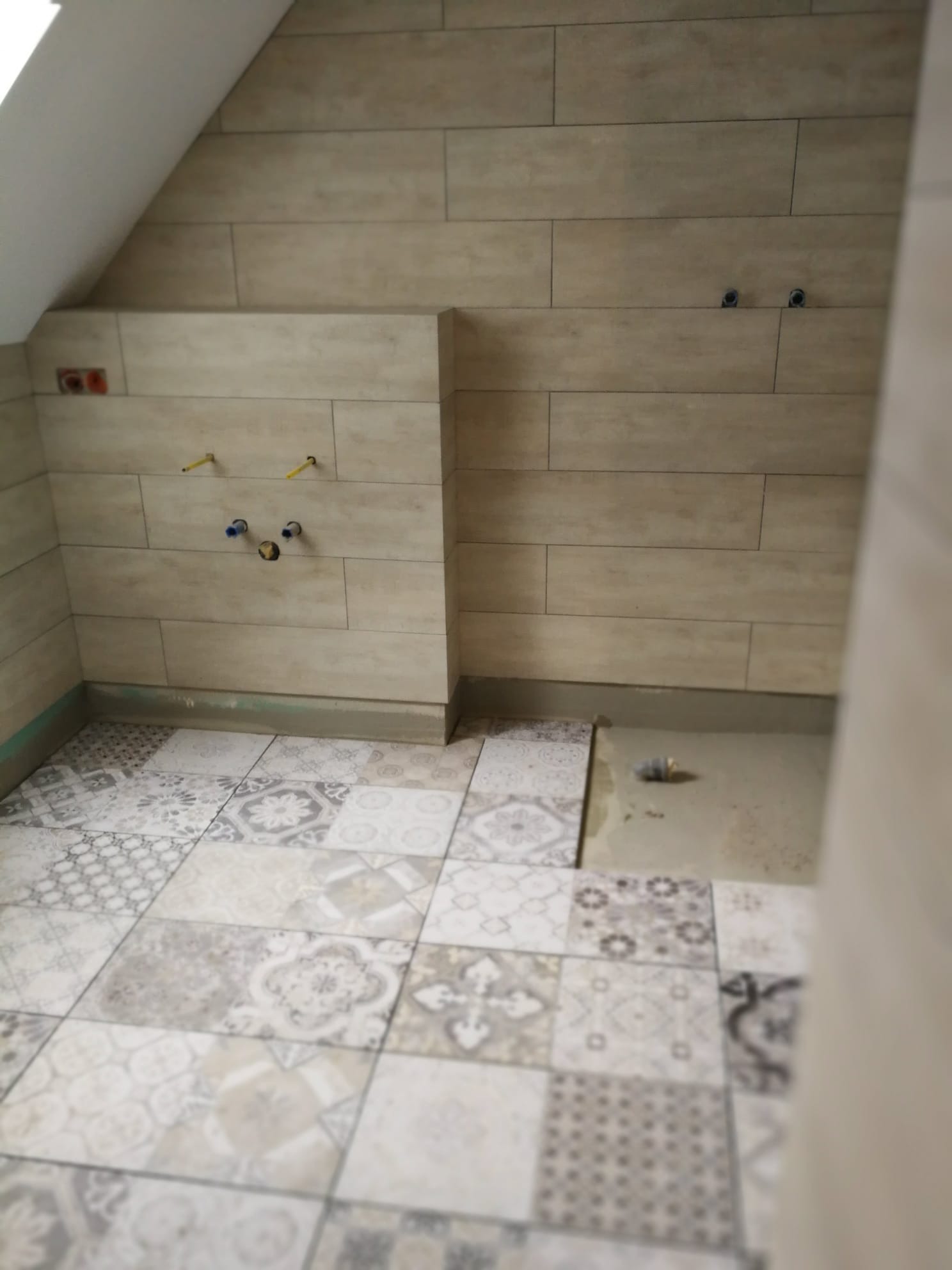 Ściana w łazience wyłożona beżowymi płytkami imitującymi drewno, z widocznymi wyprowadzonymi instalacjami wodno-kanalizacyjnymi, oraz podłoga z dekoracyjnych płytek w stylu patchwork, częściowo...