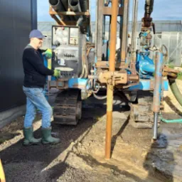 Wiertnica na gąsienicach w Żelistrzewie podczas wiercenia studni głębinowej; widoczne rury wiertnicze, operator w roboczych ubraniach i woda wypływająca z otworu.