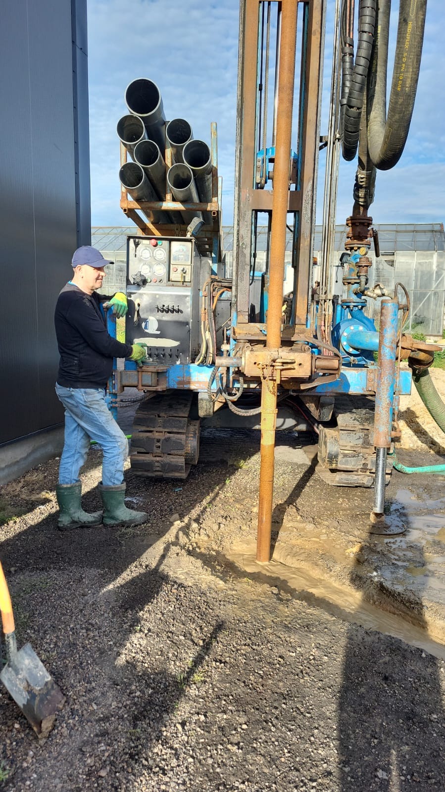 Wiertnica na gąsienicach w Żelistrzewie podczas wiercenia studni głębinowej; widoczne rury wiertnicze, operator w roboczych ubraniach i woda wypływająca z otworu.