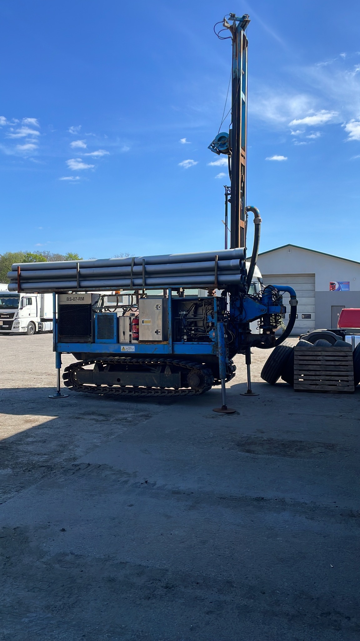 Niebieska wiertnica na gąsienicach z rurami na górze, rozstawiona na betonowym placu z białymi ciężarówkami i budynkiem w tle, błękitne niebo z chmurami.