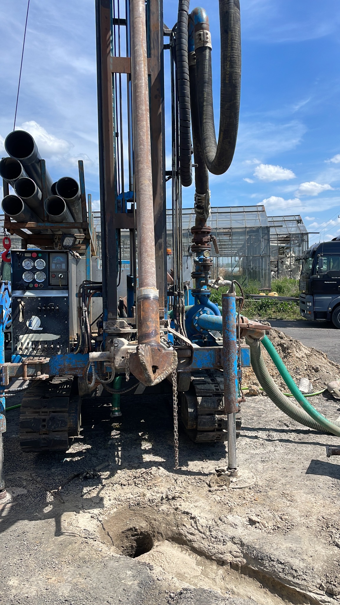 Wiertnica na gąsienicach w trakcie wiercenia otworu w ziemi, widoczne rury wiertnicze, manometry, węże, w tle szklarnie i ciężarówka.