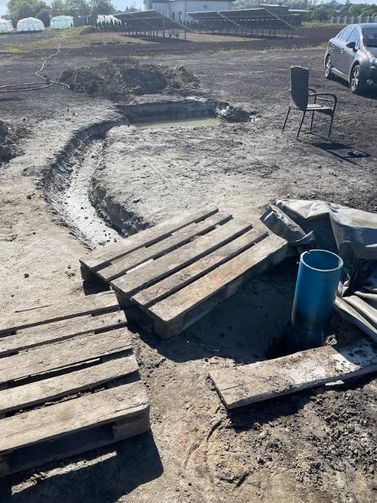 Wykop pod instalację, widoczne drewniane palety, rura PCV, wykopana ziemia i stojące krzesło ogrodowe w tle.