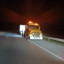 Żółta laweta z włączonymi światłami awaryjnymi stoi na pasie awaryjnym autostrady nocą, oświetlona przez reflektory i światła lawety.