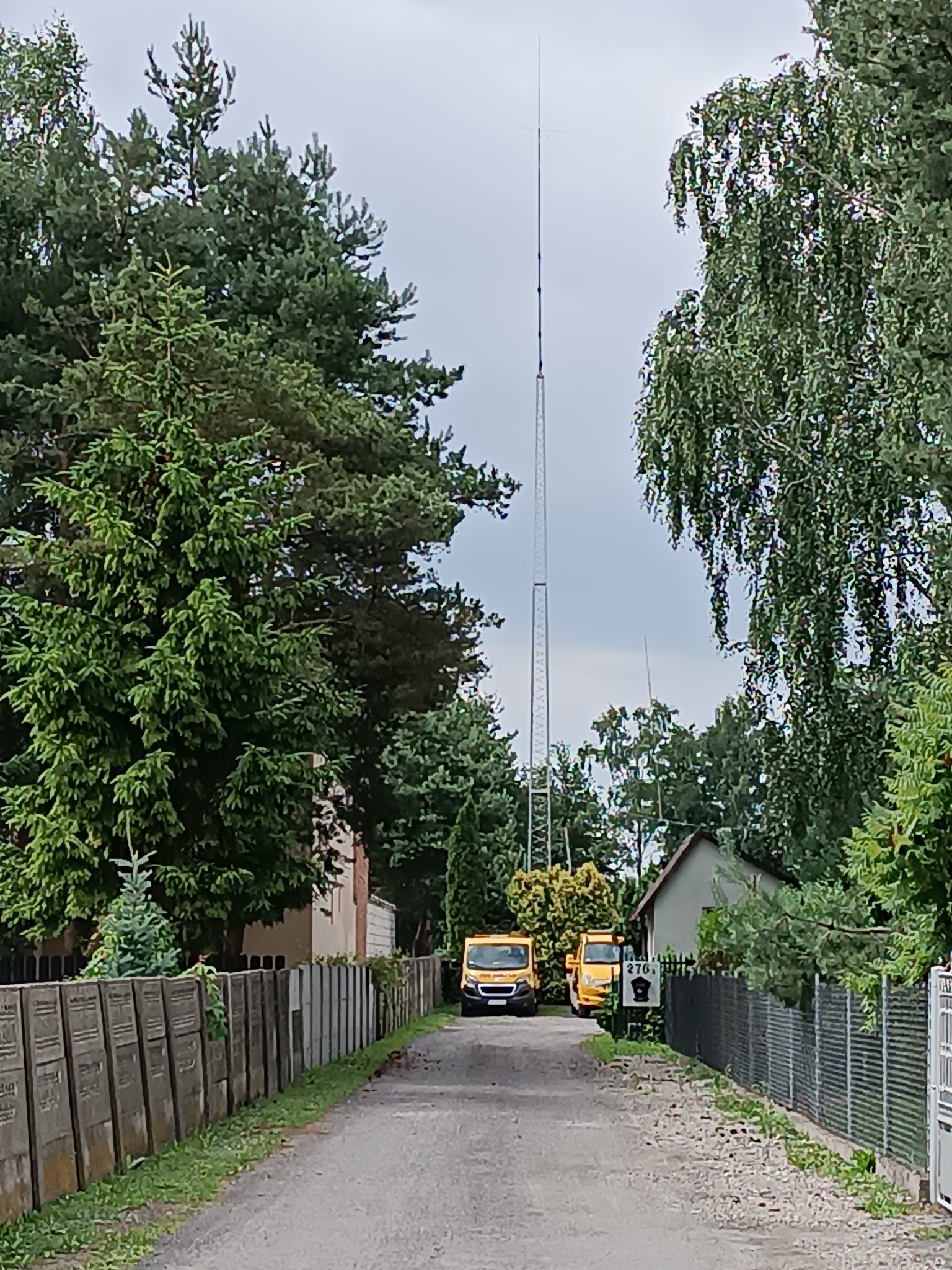 Dwa żółte samochody dostawcze zaparkowane na drodze prowadzącej do wysokiej, metalowej wieży telekomunikacyjnej, otoczonej zielenią drzew i ogrodzeniami.