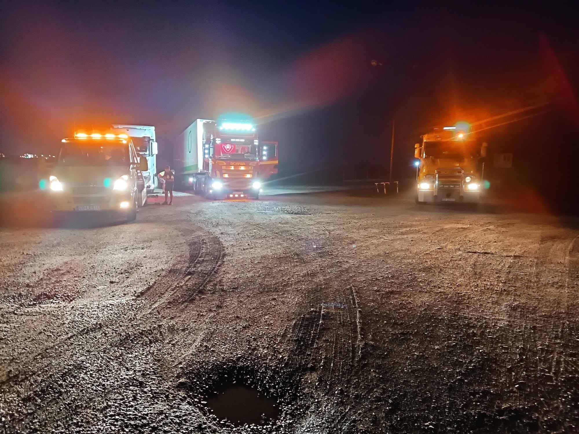 Nocne ujęcie trzech ciężarówek na nierównym terenie, z włączonymi światłami awaryjnymi i reflektorami; jedna osoba stoi obok pojazdu z lewej strony.