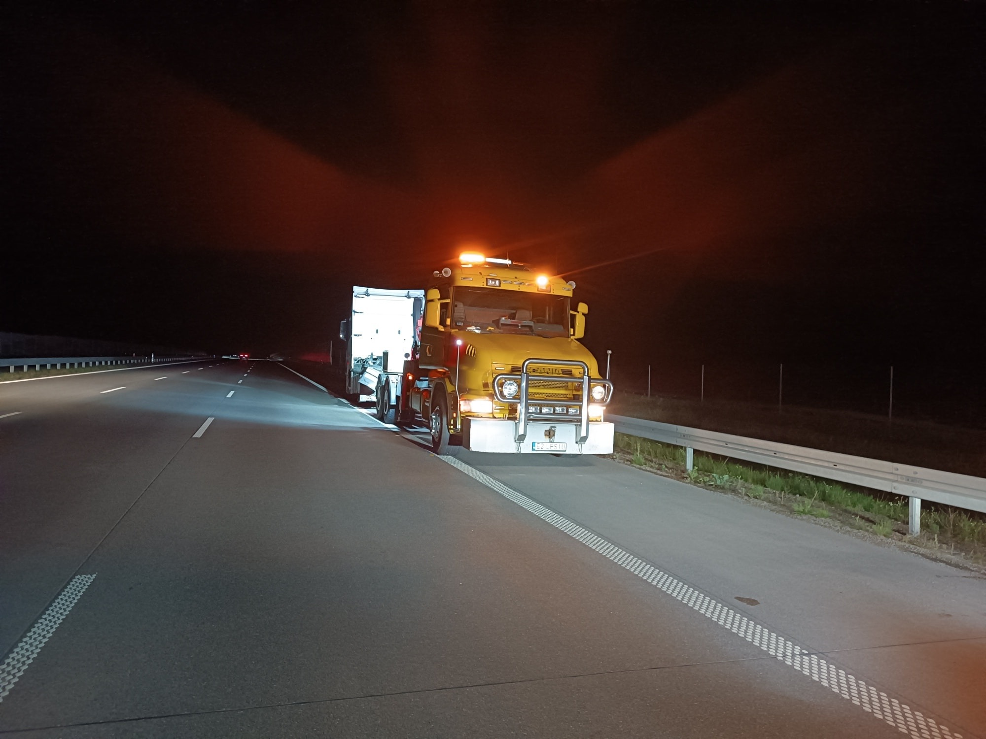 Żółta laweta z włączonymi światłami awaryjnymi stoi na pasie awaryjnym autostrady nocą, oświetlona przez reflektory i światła lawety.