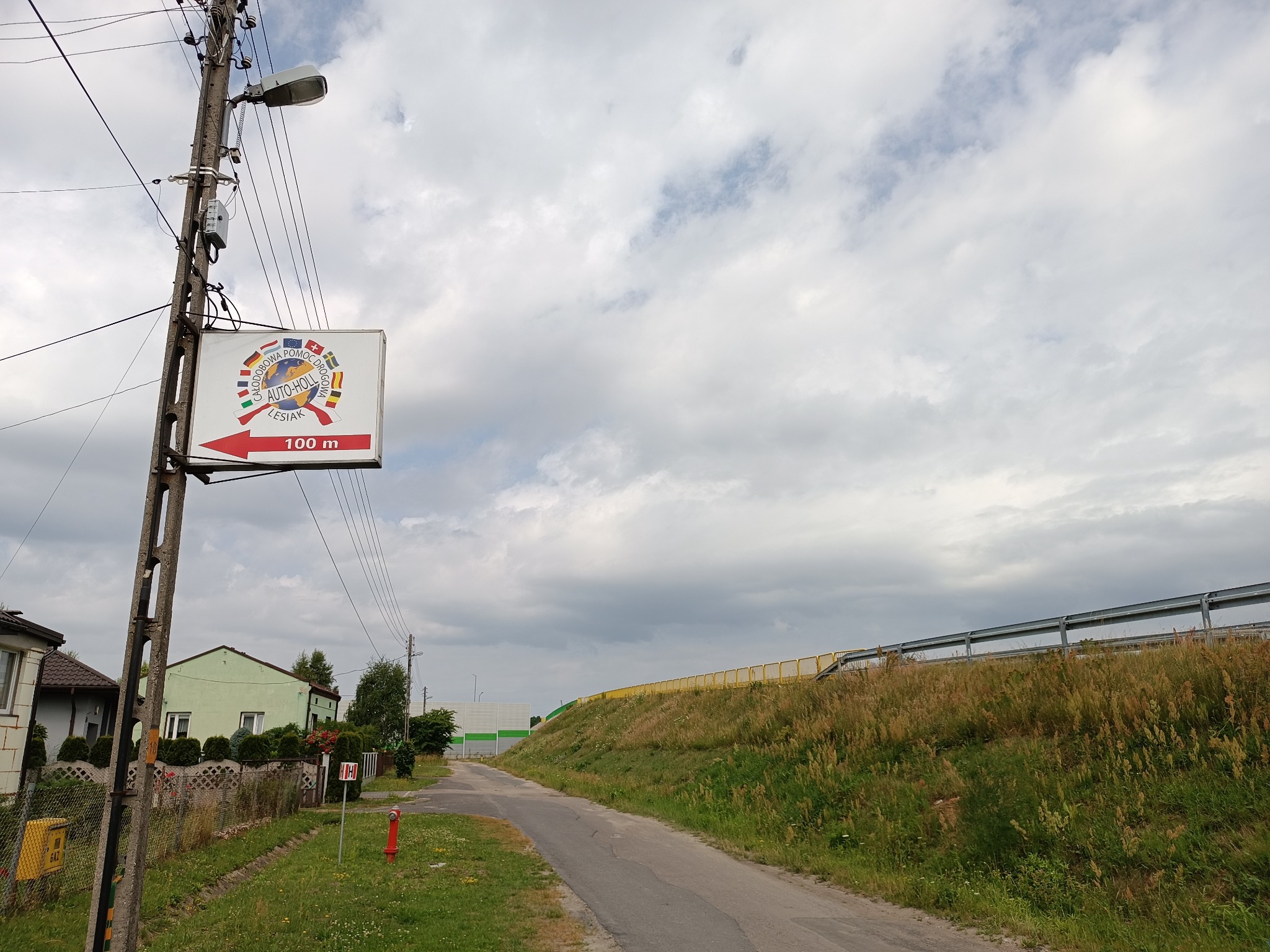 Tablica informacyjna z logo 'Całodobowa Pomoc Drogowa Auto-Holl Lesiak' na słupie energetycznym, wskazująca odległość 100m, na tle drogi i pochmurnego nieba.