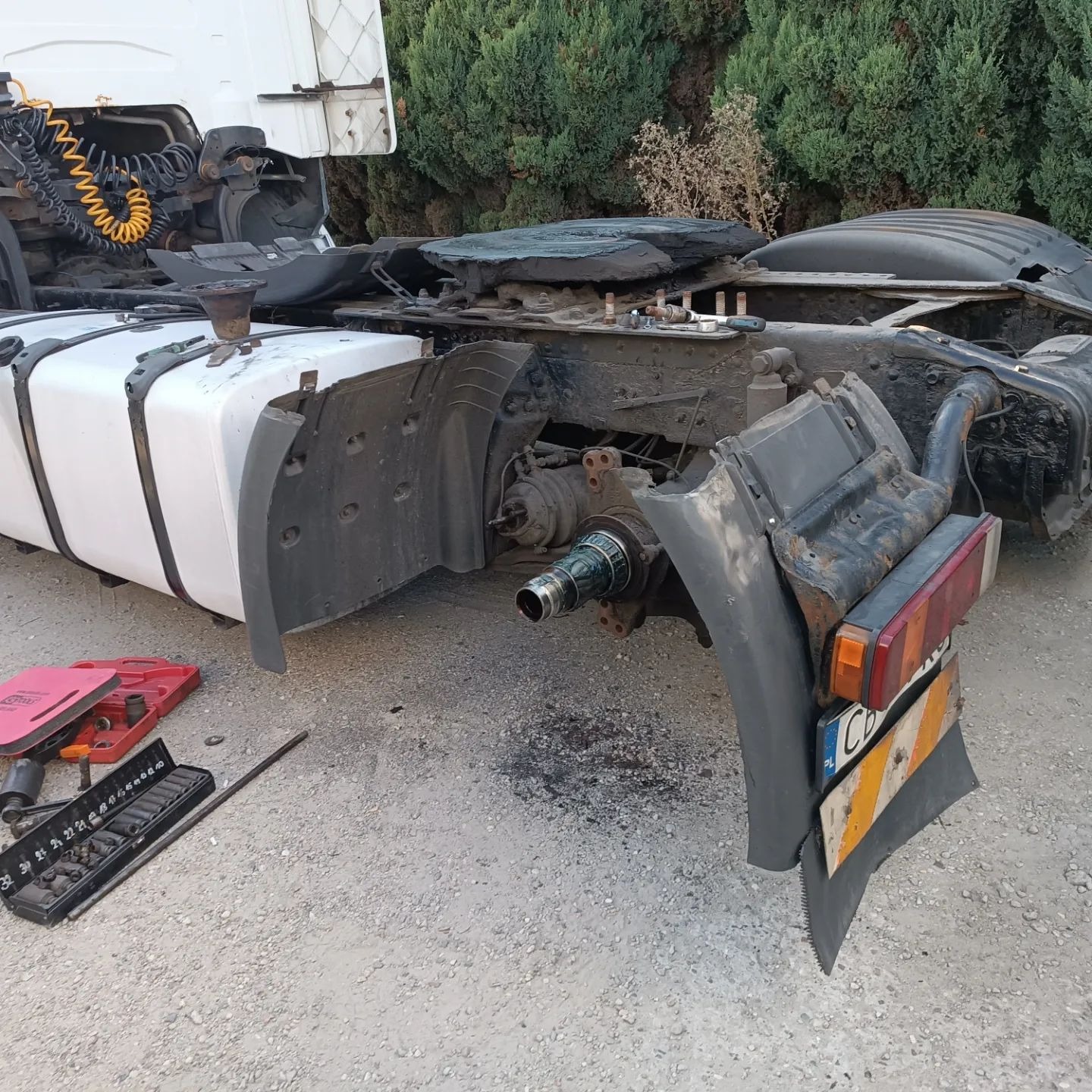 Uszkodzony samochód ciężarowy bez koła, widoczne narzędzia i elementy zawieszenia w trakcie naprawy na żwirowym podłożu.