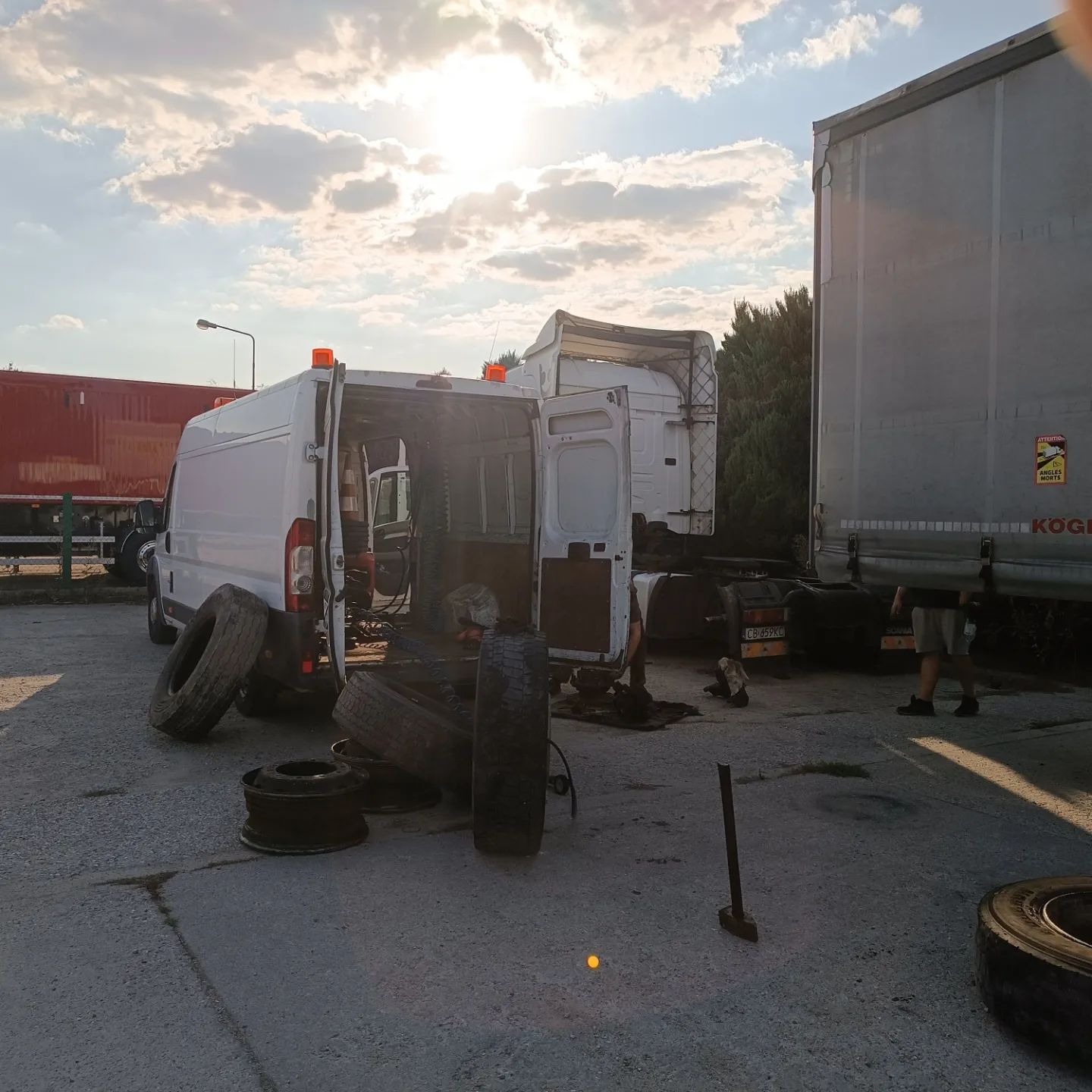 Wymiana opon w busie na parkingu, widoczne zdjęte koła, otwarte drzwi pojazdu i fragment naczepy w tle.