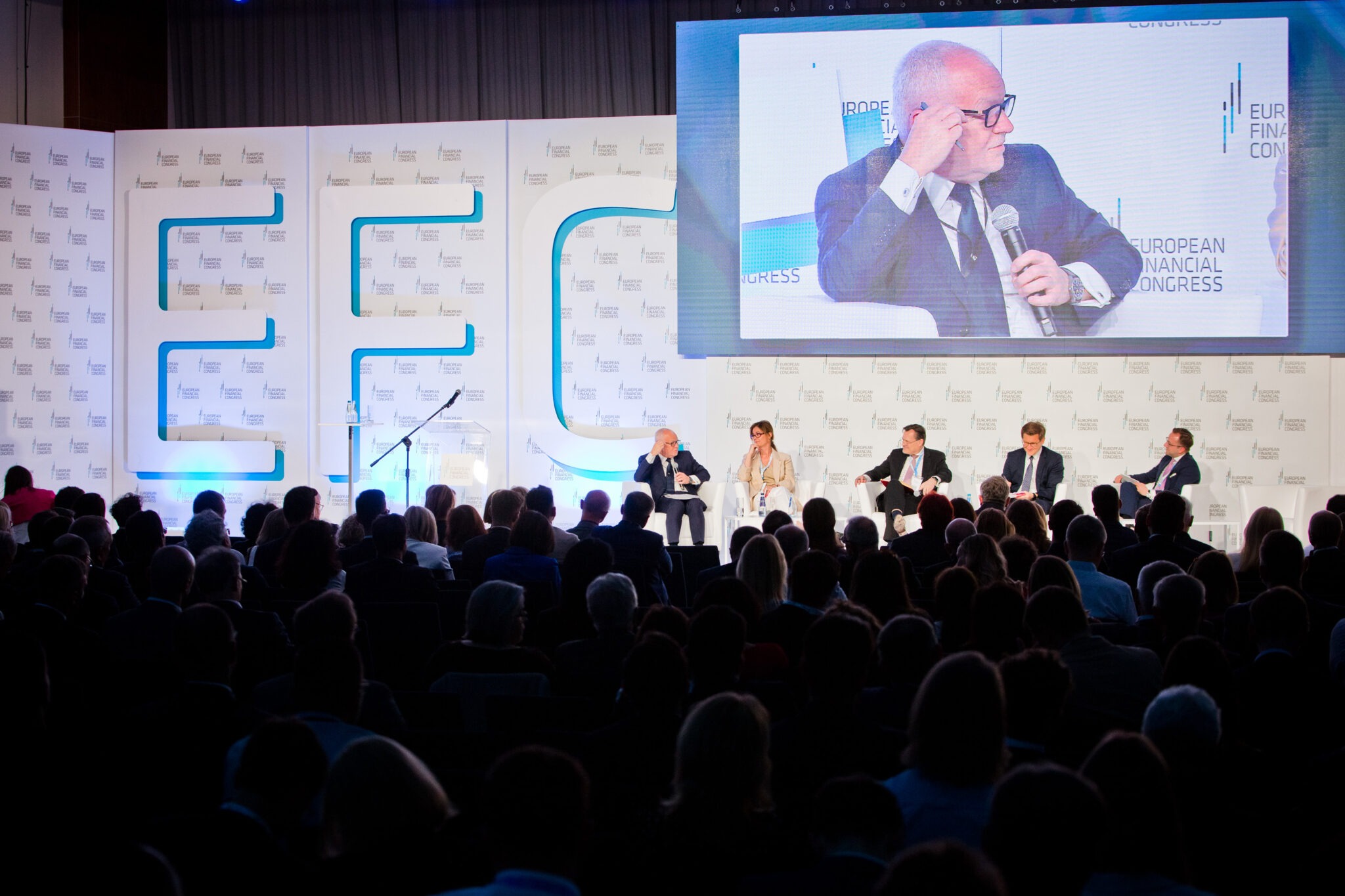 Konferencja z publicznością, panel dyskusyjny na scenie z logotypem w tle i dużym ekranem prezentującym zbliżenie mówcy.