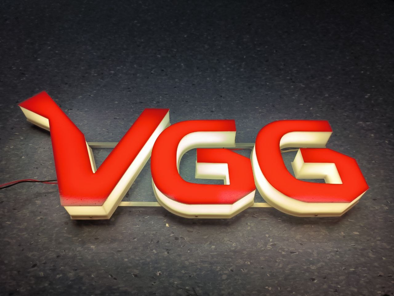 Podświetlane, czerwone litery przestrzenne VGG na szarym tle z widocznymi przewodami zasilającymi.