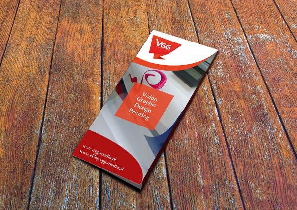 Pojedyncza, trójdzielna broszura reklamowa z logo firmy VGG, leżąca ukośnie na drewnianym blacie z widoczną fakturą drewna.