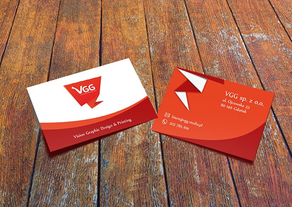Dwie wizytówki firmy VGG z czerwono-białym motywem graficznym, leżące na drewnianym stole. Jedna z logo i nazwą firmy, druga z danymi kontaktowymi.