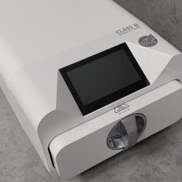 Gabinet Korona - Stomatolog | Ortodonta | Dentysta - Autoklaw stomatologiczny klasy B z wyświetlaczem, oznaczony symbolem ostrzegawczym 'gorąca powierzchnia', na szarym blacie.