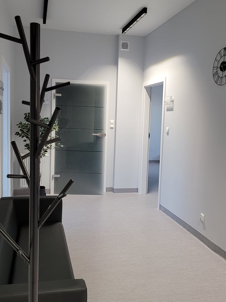 Poczekalnia w gabinecie lekarskim: czarne krzesło, wieszak w kształcie drzewa, szklane drzwi, zegar na ścianie, minimalistyczny wystrój w odcieniach szarości.