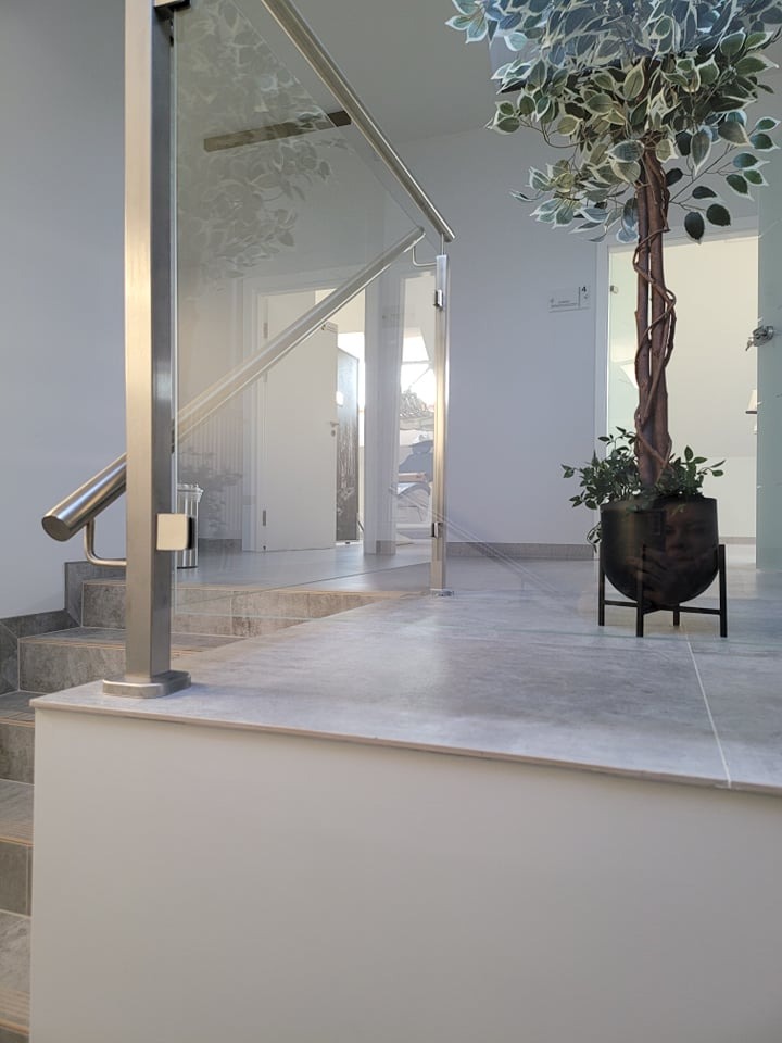 Szklana balustrada schodowa z metalowymi słupkami i poręczą, widok z dołu schodów, w tle białe ściany i drzwi, dekoracyjna roślina w donicy po prawej stronie.