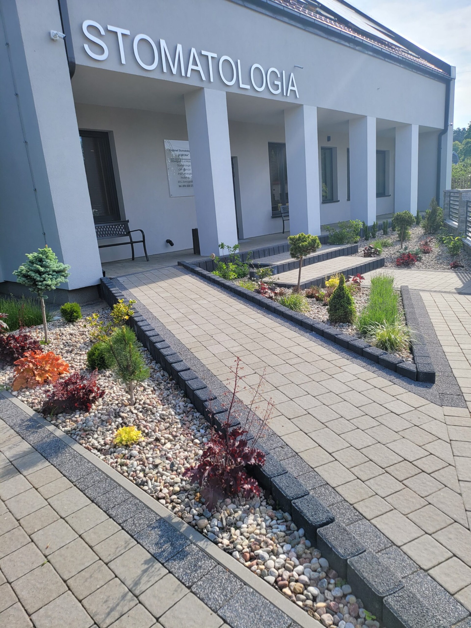 Wejście do budynku z napisem STOMATOLOGIA, ozdobny chodnik z kostki brukowej i rabaty z kamykami oraz niską zielenią.