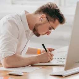 Mężczyzna w okularach, z bezprzewodowymi słuchawkami, pochylony nad szkicownikiem, rysuje czarnym cienkopisem na biurku z komputerem i klawiaturą.