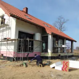 Budowa domu murowanego: robotnicy docinają styropian do izolacji fundamentów, widoczne rusztowania, ocieplenie ścian i czarna izolacja przeciwwilgociowa fundamentów.