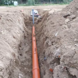 Pomarańczowa rura kanalizacyjna ułożona w wykopie w ziemi, widoczna studzienka kanalizacyjna w Mrągowie, w tle las.