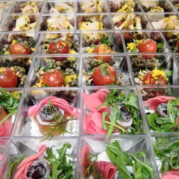 R&oacute;żnorodne przekąski w przezroczystych, kwadratowych pojemnikach: pomidorki koktajlowe z ziołami i posypką, wędlina z rukolą i marynowaną rzodkiewką oraz kawałki mięsa z sosem i przyprawami...