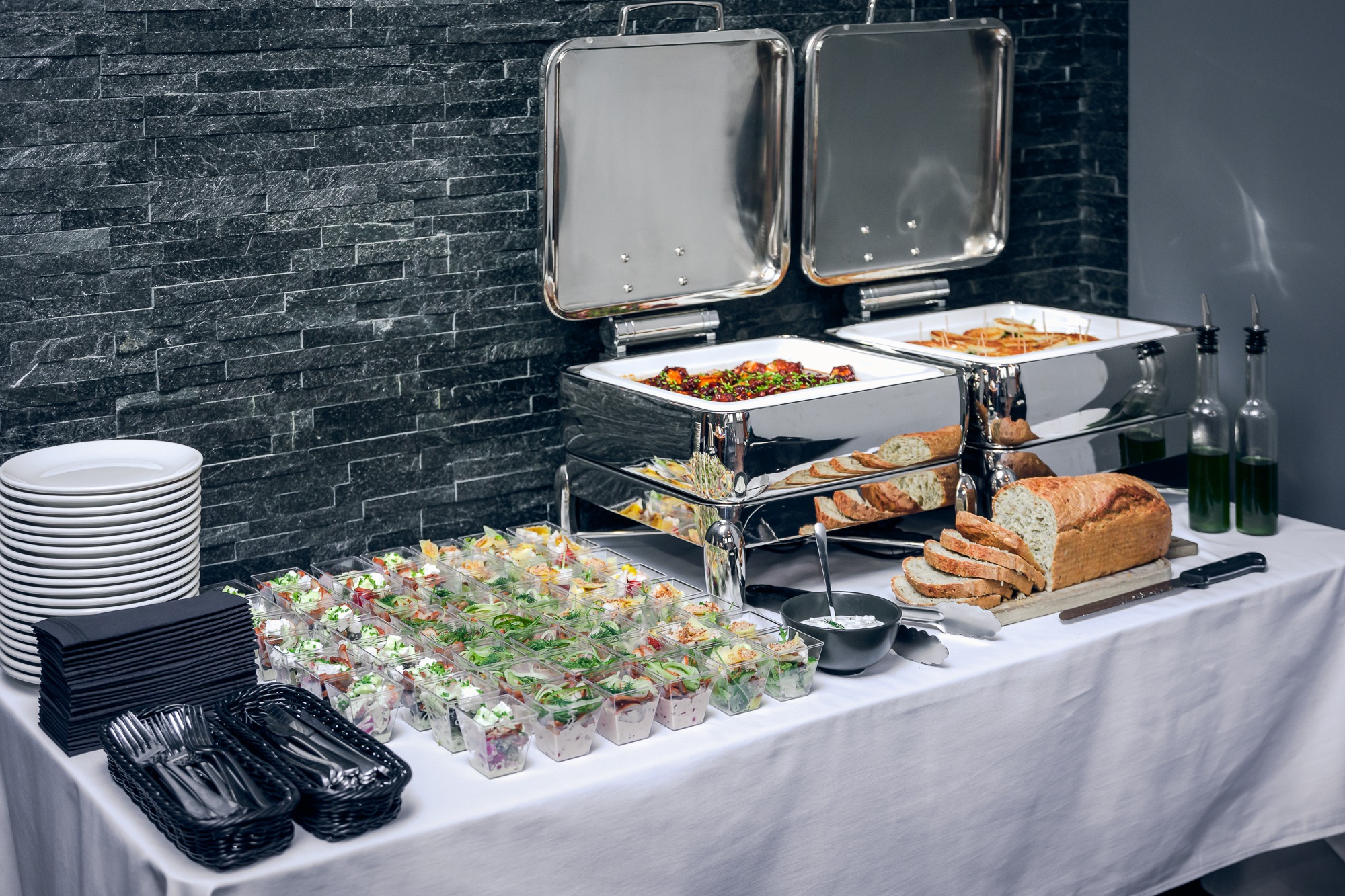 Stół cateringowy z przystawkami w plastikowych kubeczkach, chlebem na desce, podgrzewaczami z daniami oraz stosem talerzy i serwetek na tle ściany z szarego kamienia.