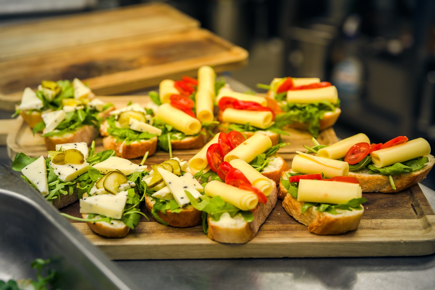 Drewniana deska z różnorodnymi kanapkami: jedne z serem pleśniowym, ogórkiem i rukolą, inne z roladkami sera żółtego i pomidorem.
