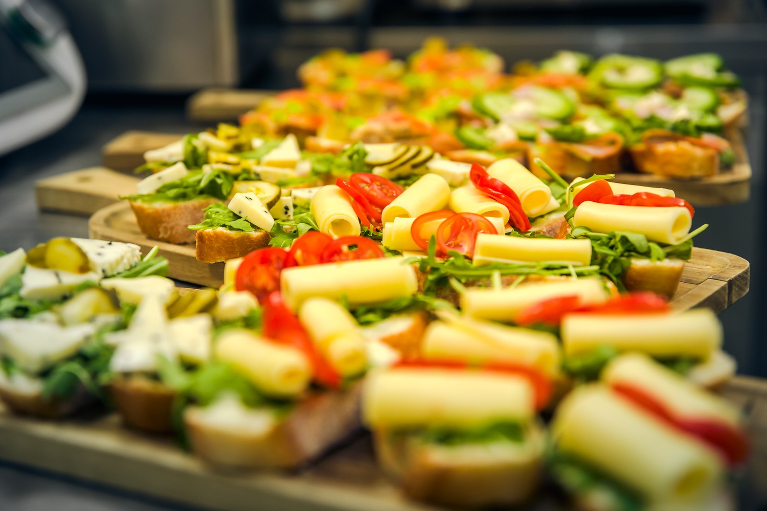 Różnorodne kanapki na drewnianych deskach: bagietki z rukolą, roladkami sera, pomidorami, ogórkami i marynowanymi warzywami, ułożone rzędami.