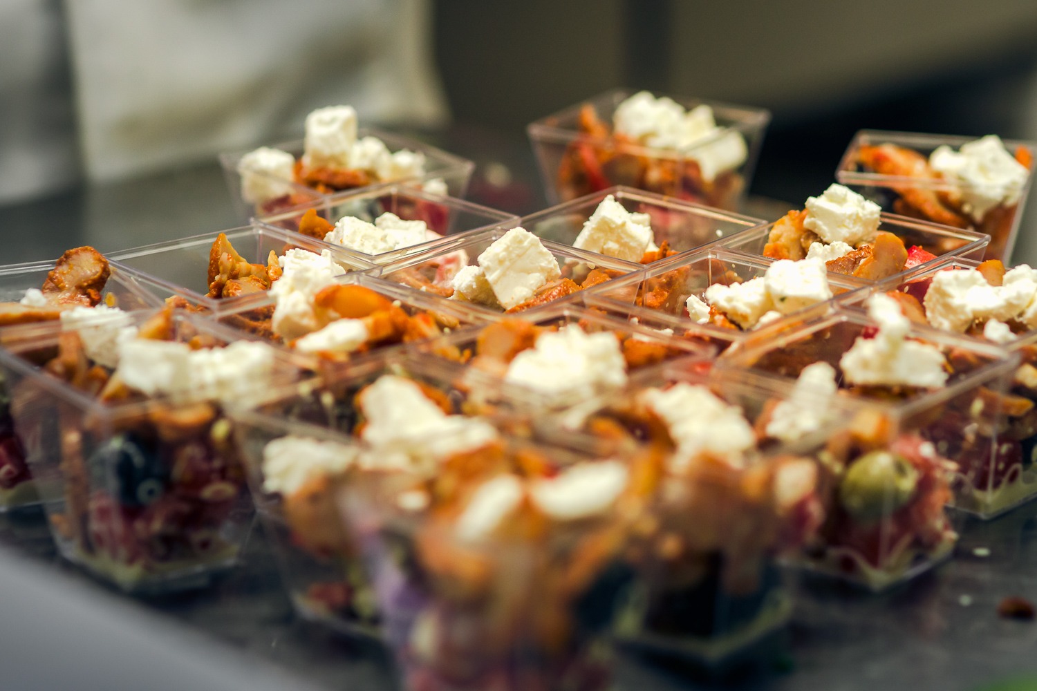 Porcje sałatek w przezroczystych, kwadratowych pojemnikach, każda z kostkami białego sera na wierzchu, ułożone rzędami na ciemnej powierzchni.